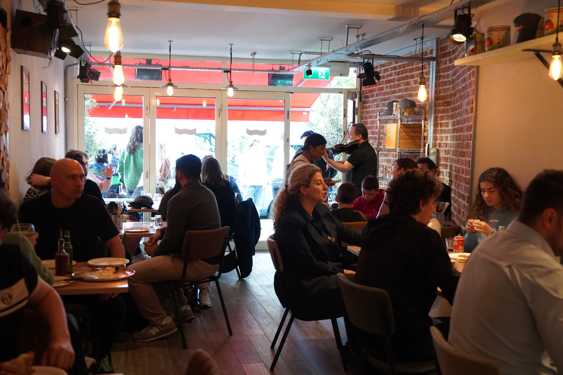 The Battersea dining room — packed, brick walls, edison bulbs and the red Pizzerium awning glowing through the window