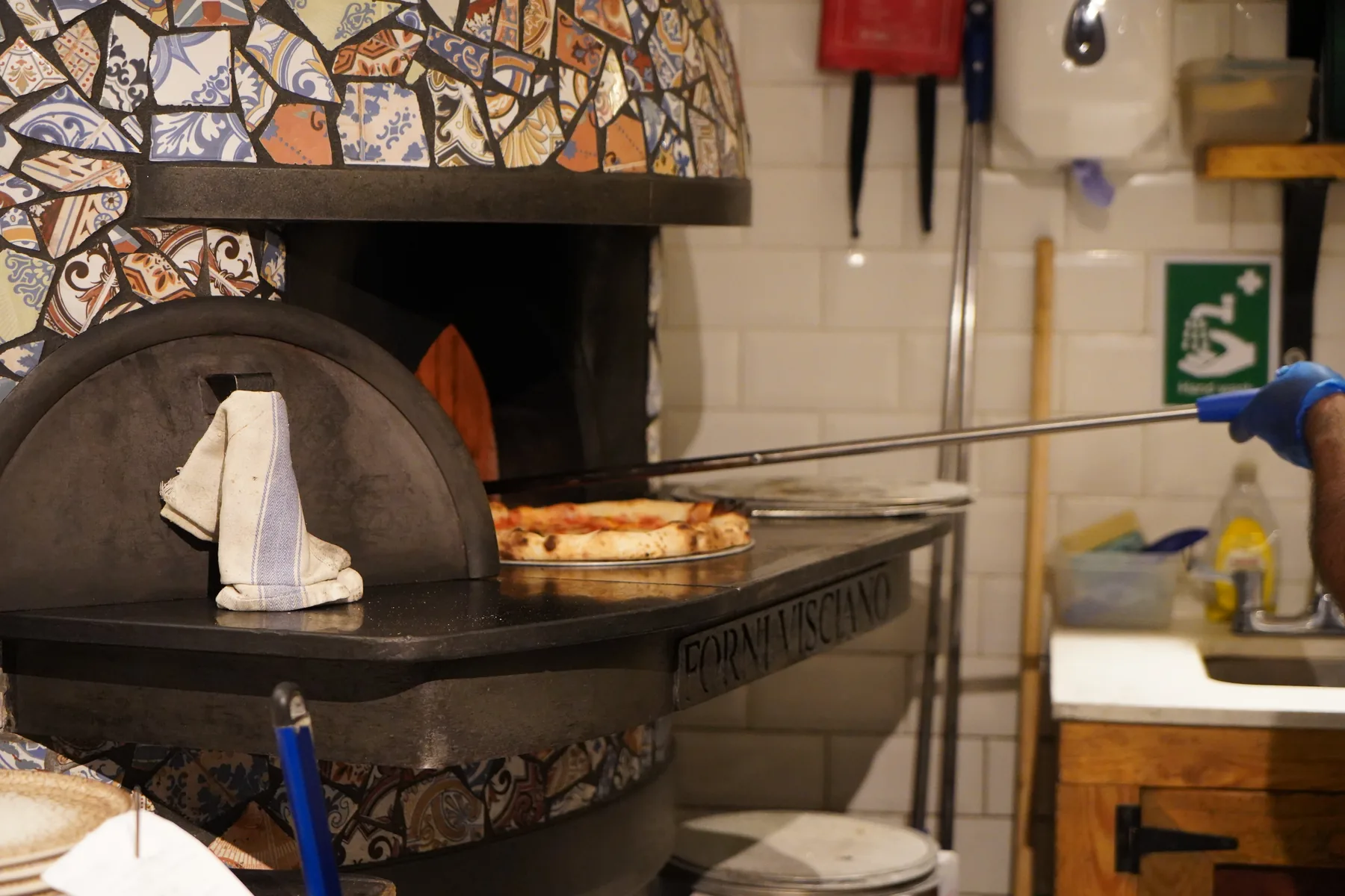 A Pizzerium pizza going into the mosaic-tiled wood-fired oven on a peel