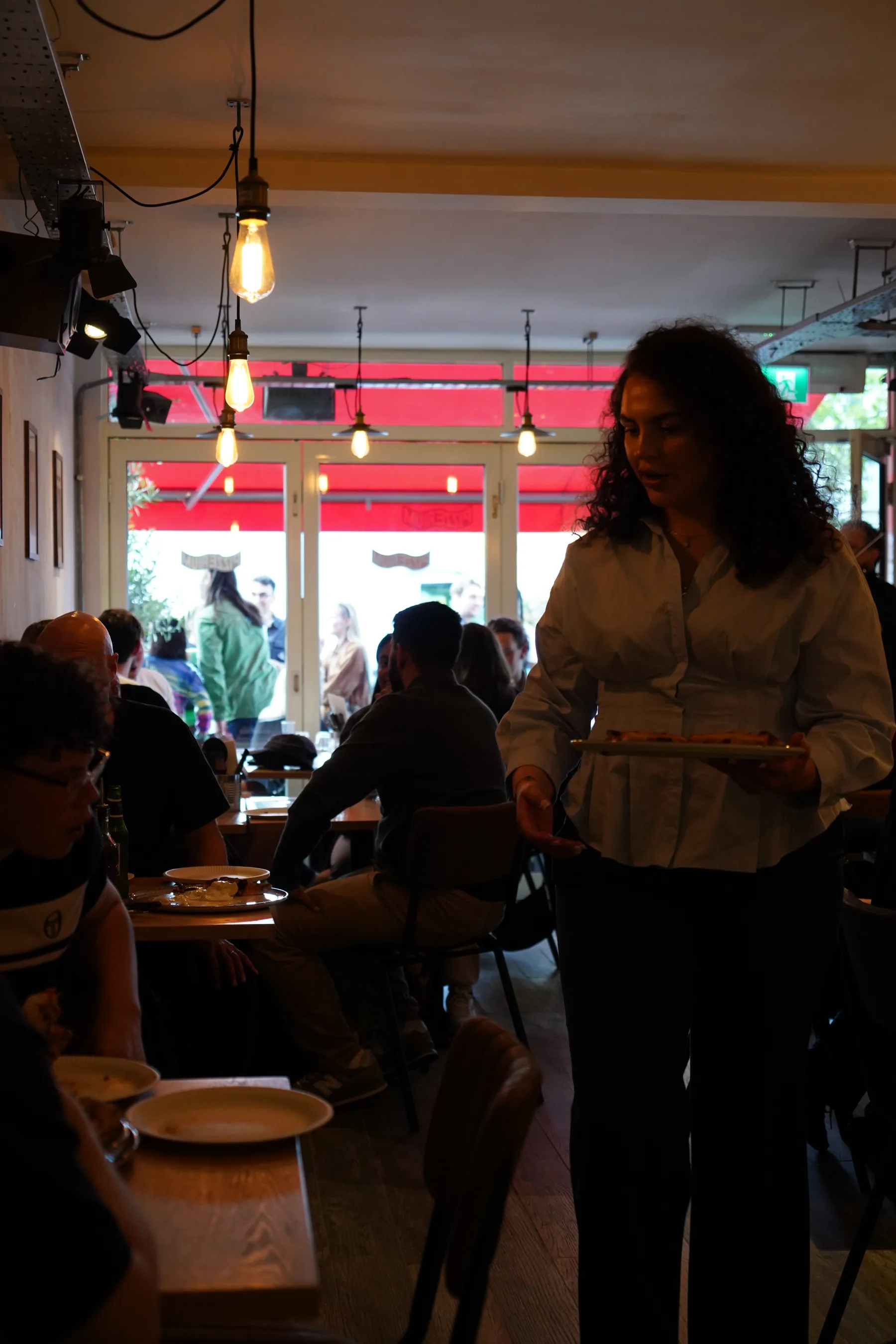 A server carrying plates through the warm-lit dining room past the red Pizzerium awning